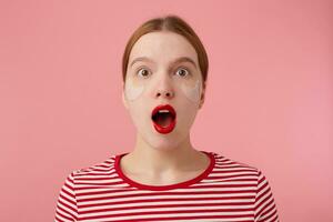 portrait de mignonne Jeune étonné roux Dame dans une rouge rayé T-shirt, avec rouge lèvres et avec patchs en dessous de le yeux, regards à le caméra avec large ouvert bouche et yeux des stands plus de rose Contexte. photo