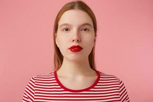 proche en haut de attrayant Jeune gingembre femme avec rad lèvres, porte dans une rouge rayé T-shirt, regards à le caméra avec une calme expression, des stands plus de rose Contexte. photo