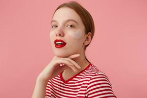 beauté souriant roux Dame porte dans une rouge rayé T-shirt, avec rouge lèvres et avec patchs en dessous de le yeux, touche le réjouissance, des stands plus de rose Contexte et profiter gratuit temps pour peau se soucier. photo