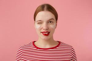 portrait de mignonne Jeune un clin d'oeil roux Dame avec rouge lèvres et avec patchs en dessous de le yeux, porte dans une rouge rayé T-shirt, regards à le caméra et mord le sien lèvres, des stands plus de rose Contexte. photo