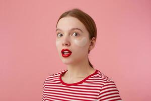 portrait de mignonne Jeune étonné roux Dame dans une rouge rayé T-shirt, avec rouge lèvres et avec patchs en dessous de le yeux, craintif regards à le caméra, des stands plus de rose Contexte. photo