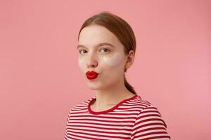 proche en haut de agréable Jeune souriant roux Dame avec rouge lèvres et avec patchs en dessous de le yeux, porte dans une rouge rayé T-shirt, regards à le caméra et envoie baiser, des stands plus de rose Contexte. photo