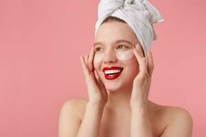 proche en haut de Jeune content femme après douche avec une serviette sur sa diriger, avec patchs et rouge lèvres, touche visage et sourires, des stands plus de rose Contexte. photo