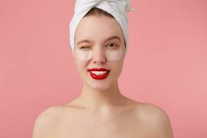 portrait de Jeune content femme après douche avec une serviette sur sa diriger, avec patchs et rouge lèvres, à la recherche à le caméra et un clin d'oeil plus de rose Contexte. photo