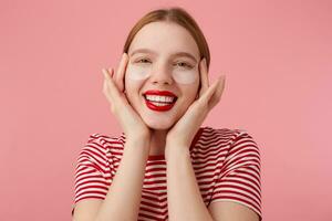 agréable Jeune roux Dame dans une rouge rayé T-shirt, avec rouge lèvres , touche le sien visage avec des doigts, très heureux avec mon Nouveau patchs de foncé cercles en dessous de mon yeux. des stands plus de rose Contexte. photo