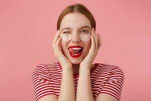 portrait de mignonne Jeune un clin d'oeil roux Dame avec patchs en dessous de le yeux, avec rouge lèvres et , porte dans une rouge rayé T-shirt, regards à le caméra et spectacles langue , des stands plus de rose Contexte. photo