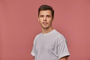 intérieur fermer de attrayant Jeune foncé aux cheveux Masculin avec court la Coupe de cheveux portant décontractée vêtements, permanent plus de rose Contexte avec mains bas, à la recherche à caméra avec calme visage photo