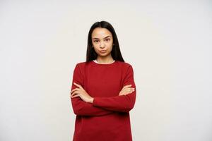 portrait de sérieux, adulte asiatique fille avec foncé longue cheveux. portant rouge chandail et plis franchi bras sur une poitrine. en train de regarder à le caméra réprobateur, isolé plus de blanc Contexte photo