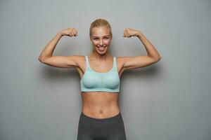 jolie sportif Jeune longue aux cheveux blond femme élevage mains et montrant sa pouvoir, menthe Haut photo