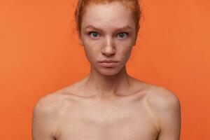 intérieur coup de jolie Jeune tête de lecture femelle avec décontractée coiffure et non maquillage permanent plus de Orange arrière-plan, à la recherche à caméra avec perplexe affronter, arrondi yeux étonné avec plié lèvres photo