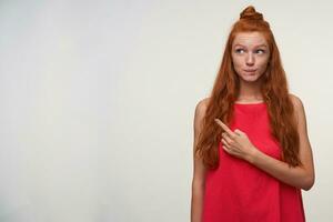 rêveur charmant Jeune tête de lecture Dame avec ondulé cheveux portant non faire en haut, permanent plus de blanc Contexte dans décontractée robe, montrant de côté avec indice doigt, élevage les sourcils et mordant lèvre inférieure photo