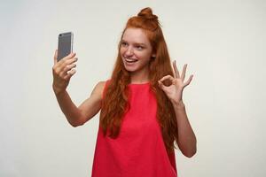 portrait de charmant Jeune Dame dans rose robe portant sa rusé cheveux dans chignon, fabrication photo de elle-même avec mobile téléphone, souriant largement à caméra et rasant main avec D'accord geste