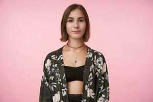 intérieur photo de Jeune charmant court aux cheveux brunette femelle avec Naturel maquillage à la recherche positivement à caméra avec lumière sourire tandis que posant plus de rose Contexte