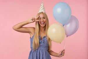 marrant intérieur coup de Jeune blond femme avec longue cheveux donnant clin d'œil et élevage D'accord geste Heureusement, en portant multicolore air des ballons dans sa mains tandis que posant plus de rose Contexte photo