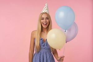 content Jeune longue aux cheveux femelle posant dans multicolore air des ballons, portant bleu été robe et anniversaire casquette, célébrer vacances, isolé plus de rose Contexte photo