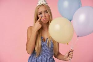 dérangé longue aux cheveux blond femme dans posant dans bleu été robe et anniversaire casquette cône chapeau en portant multicolore air des ballons, à la recherche à caméra Malheureusement essuyage larmes loin, isolé plus de rose Contexte photo