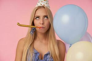 ennuyé jolie blond femme avec longue cheveux roulant yeux et en gardant fête klaxon dans sa bouche, célébrer anniversaire avec multicolore air des ballons, étant mécontent avec faire la fête, posant plus de rose Contexte photo