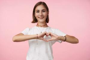 charmant Jeune de bonne humeur court aux cheveux brunette femelle avec Naturel maquillage formant cœur avec élevé mains et souriant largement à caméra, permanent plus de rose Contexte photo