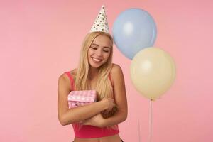 portrait de attrayant Jeune blond femelle avec longue cheveux en gardant cadeau boîte et souriant Cordialement avec fermé yeux, montrant sa agréable émotions plus de rose Contexte photo