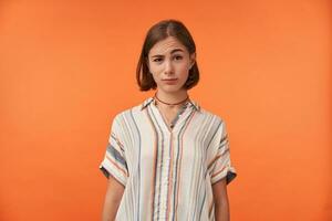 portrait de Jeune isolé femme contre Orange Contexte à la recherche à vous avec un sourcil levé, portant rayé chemise. lorsque vous entendre quelque chose bizarre. photo