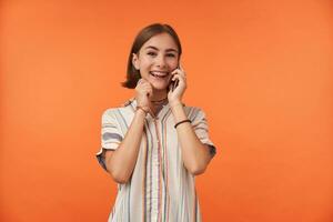 femelle étudiant parler sur intelligent téléphone, portant rayé chemise, les dents un appareil dentaire et bracelets, entendre une bien nouvelles. souriant fille à la recherche content et en train de regarder à le caméra isolé plus de Orange Contexte photo