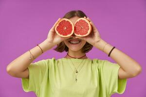 femelle étudiant avec gros sourire, Jeune Dame avec court brunette cheveux en portant pamplemousse plus de sa yeux. permanent plus de violet Contexte. portant vert T-shirt, les dents un appareil dentaire et bracelets photo