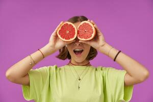 adolescent fille, de bonne humeur et content, femme avec brunette court cheveux en portant pamplemousse plus de sa yeux. permanent plus de violet Contexte. portant vert T-shirt, les dents croisillons, bracelets et Collier photo