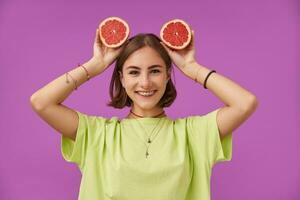 adolescent fille, de bonne humeur et content souriant Dame avec court brunette cheveux en portant pamplemousse plus de sa diriger. permanent plus de violet Contexte. portant vert T-shirt, les dents croisillons, bracelets et Collier photo