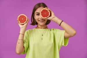 portrait de attractif, mignonne fille avec court brunette cheveux. en portant pamplemousse plus de sa œil, couverture un œil. permanent plus de violet Contexte. portant vert T-shirt, collier, un appareil dentaire et bracelets photo