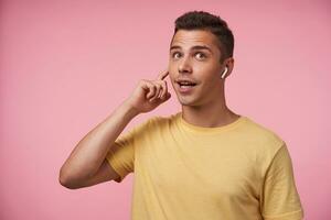 excité Jeune attrayant court aux cheveux homme habillé dans Jaune T-shirt à la recherche émotionnellement vers le haut tandis que ayant téléphone appel avec casque, permanent plus de rose Contexte photo