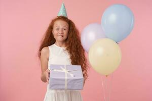 fou de joie frisé femelle enfant avec longue rusé cheveux posant plus de rose Contexte avec cadeau emballé boîte et à la recherche à caméra gaiement, portant blanc élégant robe et anniversaire casquette photo