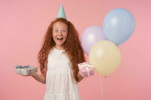 joyeux femelle frisé enfant avec longue rusé cheveux étant excité et surpris à avoir anniversaire cadeau, Heureusement à la recherche dans caméra avec cadeau boîte dans mains plus de rose Contexte. les enfants et fête concept photo