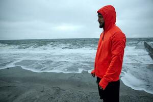 horizontal coup de Jeune foncé aux cheveux barbu sportif habillé dans chaud Orange sweat à capuche et noir athlétique pantalon permanent plus de bord de mer sur gris de bonne heure Matin et à la recherche pensivement devant photo