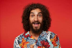 studio photo de de bonne humeur jolie Jeune frisé barbu Masculin avec frisé marron cheveux permanent contre rouge Contexte dans chemise avec floral imprimer, à la recherche Heureusement à caméra avec large bouche ouvert