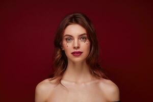 portrait de magnifique aux yeux bleus Jeune brunette femme avec de fête maquillage et argent étoiles sur sa affronter, à la recherche calmement à caméra tandis que permanent plus de bordeaux Contexte photo