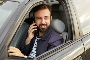 photo de de Jeune attrayant réussi barbu homme dans une bleu veste et rayé T-shirt, est assis derrière le roue de le voiture et attend pour une réponse sur le téléphone.