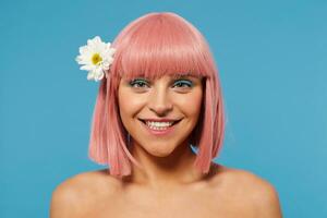 portrait de Jeune charmant positif rose aux cheveux femelle avec court la Coupe de cheveux à la recherche Heureusement à caméra avec charmant sourire, ayant camomille dans sa cheveux tandis que permanent plus de bleu Contexte photo