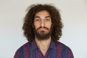 portrait de Jeune Beau aux yeux bruns barbu Masculin avec foncé frisé cheveux à la recherche à caméra avec calme affronter, en gardant lèvres plié tandis que permanent contre blanc Contexte photo