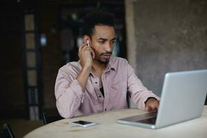 barbu homme avec foncé peau écoute à conférences avec écouteurs avant examen, en utilisant le sien portable et Publique Wifi dans ville café, à la recherche à écran avec sérieux visage et en portant main sur clavier photo