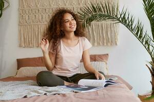 photo de rêveusement Jeune africain américain femme avec frisé cheveux, est assis sur le lit et regards loin, sourit et lit une Nouveau Voyage magazine.