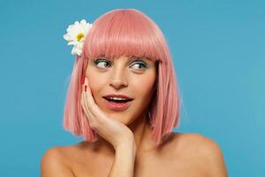 fermer de Jeune charmant femelle avec court rose la Coupe de cheveux penché sa menton sur élevé main tandis que à la recherche positivement de côté avec lumière sourire, isolé plus de bleu Contexte photo