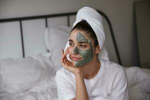 portrait de attrayant Jeune foncé aux cheveux femme avec serviette sur sa tête penché menton sur élevé main et à la recherche de côté pensivement tandis que en gardant masque sur sa affronter, séance chambre intérieur photo