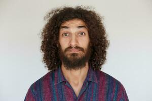 studio photo de attrayant aux yeux bruns Jeune barbu Masculin avec foncé frisé cheveux élevage les sourcils avec surprise tandis que à la recherche à caméra, habillé dans rayé multicolore chemise contre blanc Contexte