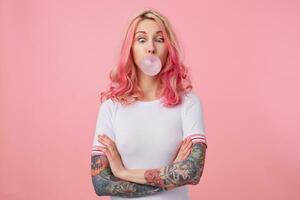 Jeune marrant magnifique rose aux cheveux femme avec tatoué mains, porte dans blanc T-shirt, coups gencive balle, plissé yeux sur nez, des stands plus de rose Contexte avec franchi bras. photo