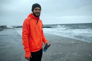 coup de Jeune de bonne humeur barbu sportif Masculin avec sourcil perçant en portant bouteille avec l'eau dans le sien main tandis que permanent plus de littoral de mer sur orageux du froid temps, souriant positivement à caméra photo