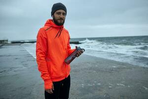 Jeune attrayant barbu sportif habillé dans chaud athlétique vêtements en marchant le long de front de mer après fonctionnement session, écoute à la musique avec écouteurs et souriant positivement à caméra photo