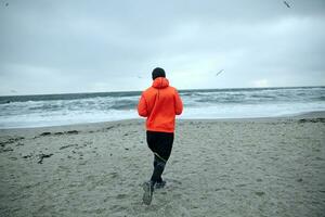 retour vue de aptitude Masculin modèle travail en dehors et fonctionnement par bord de mer sur gris de bonne heure matin, habillé dans chaud sportif vêtements et Orange manteau. sport et en bonne santé mode de vie concept photo