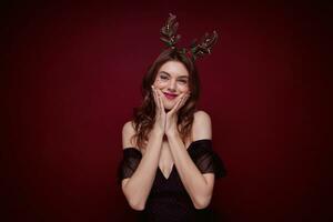 studio photo de Jeune marron aux cheveux attrayant femelle portant soir maquillage pendant chrismas faire la fête, en portant sa des joues avec élevé paumes et ayant amusement plus de bordeaux Contexte