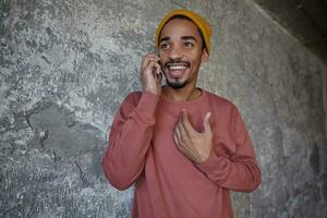 bien à la recherche jolie Jeune foncé écorché homme avec marron yeux ayant agréable conversation avec le sien téléphone intelligent, étant dans agréable ambiance et souriant gaiement plus de béton Contexte photo