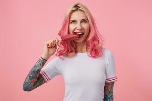 portrait de Jeune magnifique aux cheveux roses fille avec tatoué mains, portant une blanc T-shirt, à la recherche à le caméra et en essayant à mordre de le la glace crème, permanent plus de rose Contexte. photo